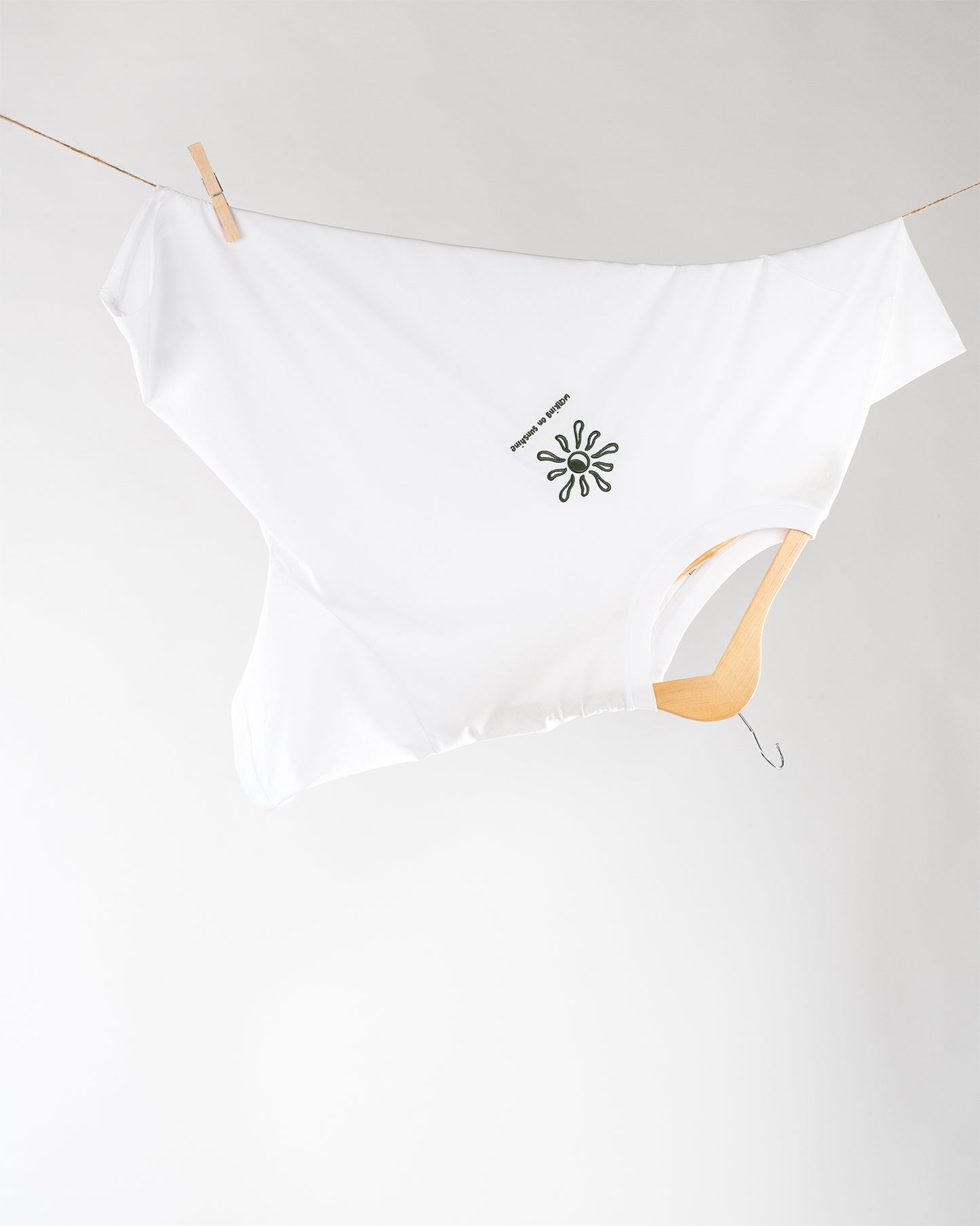 Weißes T-Shirt, aufgehängt auf einem Holzkleiderbügel und einer Wäscheleine, seitlich fixiert mit einer Holzklammer. In der Mitte des T-Shirts befinmdet sich eine Sonnenform mit dem Text ‚WALKING ON SUNSHINE‘.
