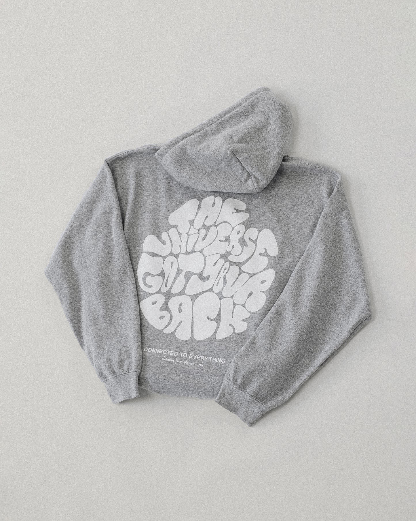 Flach liegender grauer Kapuzenpullover, Rückseite sichtbar. Auf dem Rücken ist ein großer weißer Print mit dem Schriftzug „THE UNIVERSE GOT YOUR BACK“ in organisch geschwungenen Großbuchstaben. Darunter in kleinerer Schrift: „CONNECTED TO EVERYTHING – clothing from planet earth“.