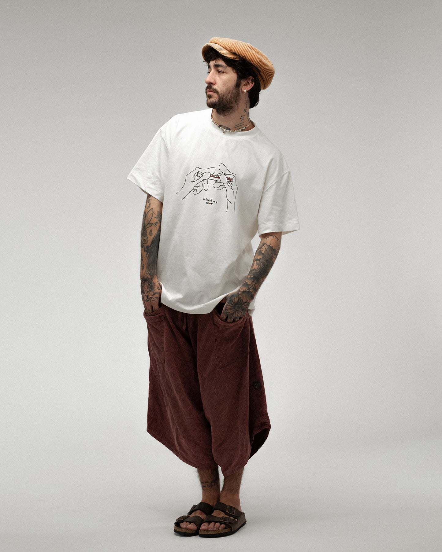Modell trägt ein weißes T-Shirt mit schwarz-rote Liniezeichnung zweier Hände, die einen Joint rollen. In der Mitte des Motivs der Schriftzug „inhale my love“.