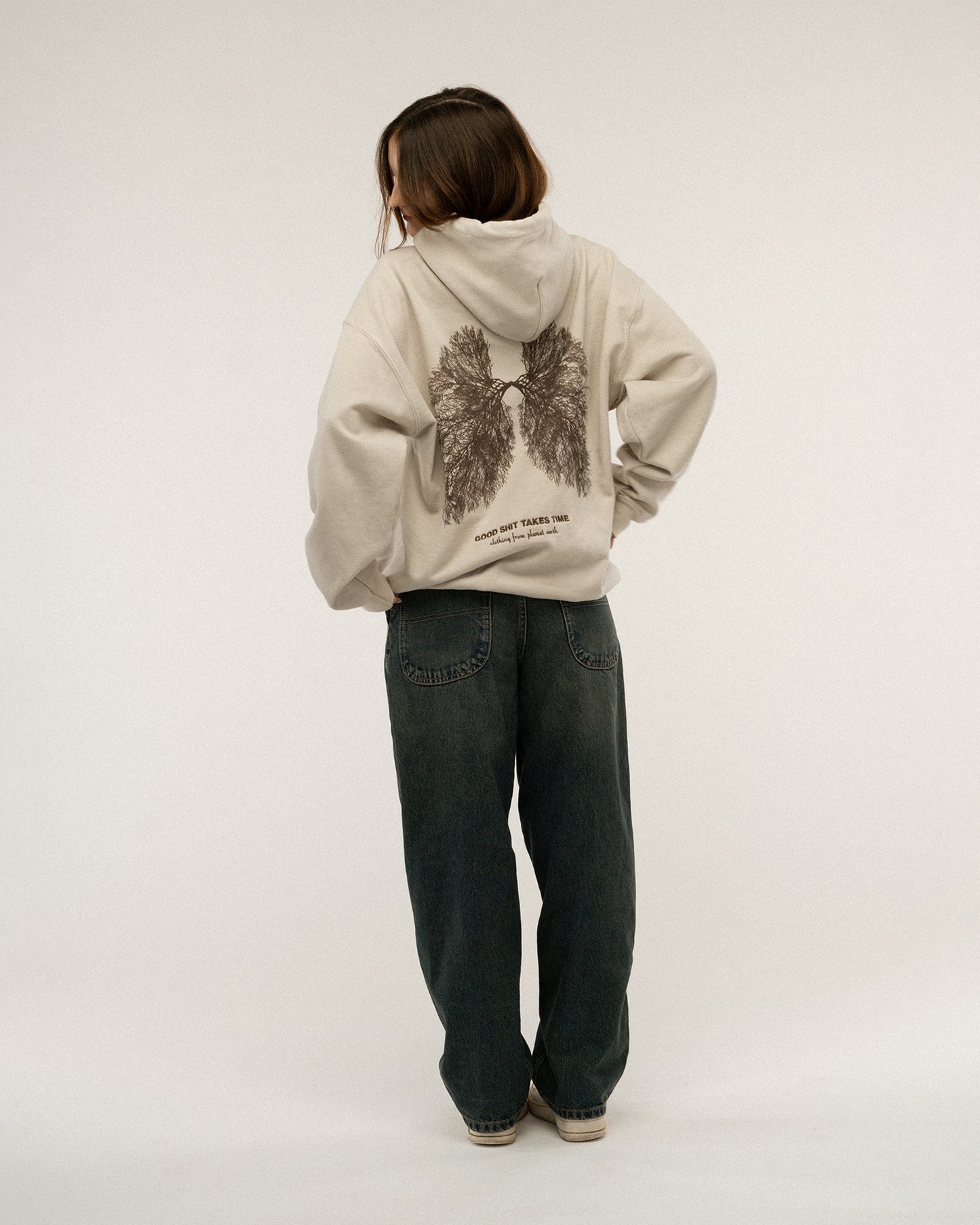 Beiger Kapuzenpullover mit großem dunkelbraunem Print auf dem Rücken. Die Illustration zeigt Lungenflügel aus feinen Baumwurzeln mit dem Text „GOOD SHIT TAKES TIME“ darunter. Das Model steht ruhig mit Blick zur Seite.