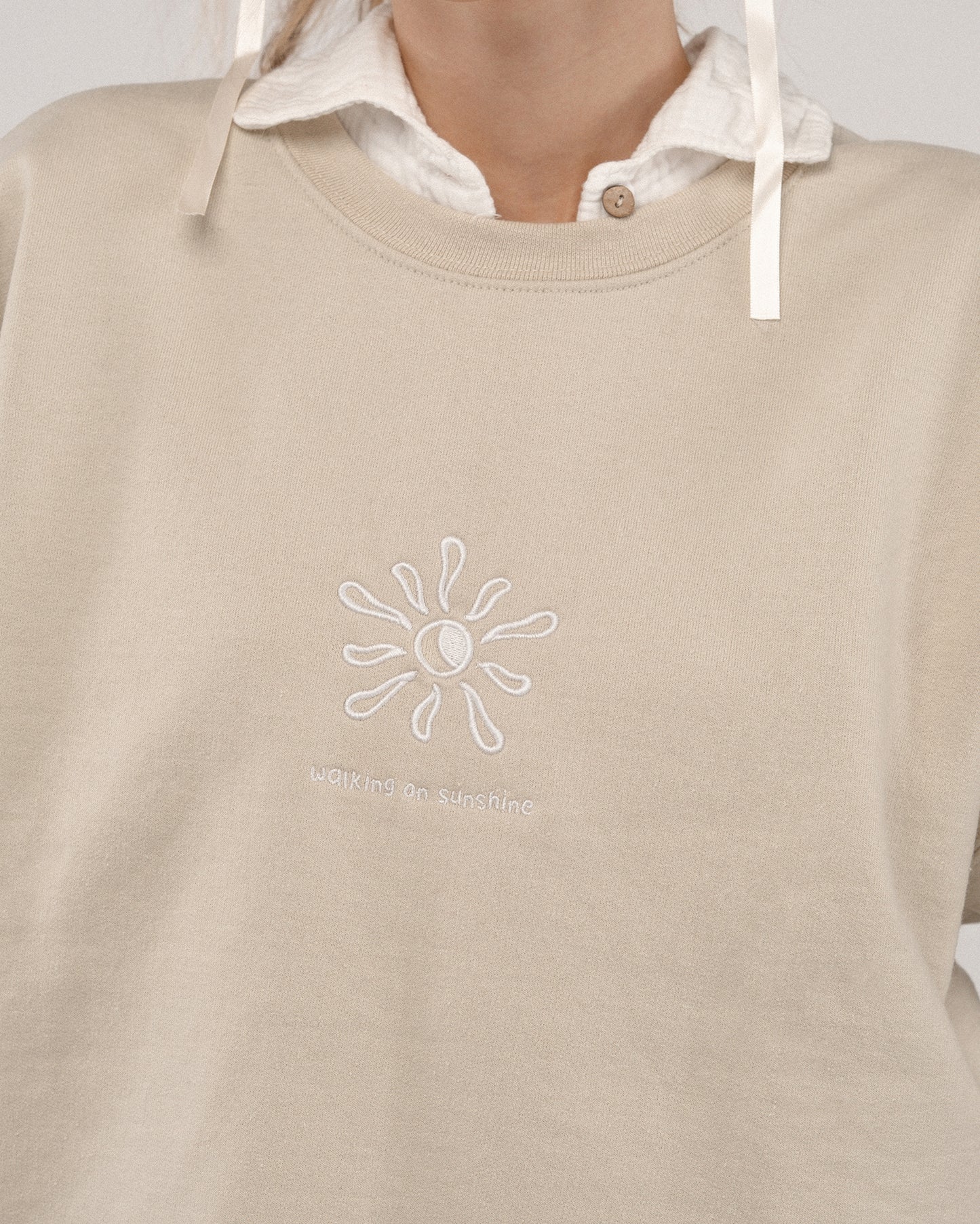 Nahaufnahme der Brustpartie eines beigefarbenen Sweatshirts mit weißem Sonnenmotiv und dem gestickten Schriftzug „walking on sunshine“.