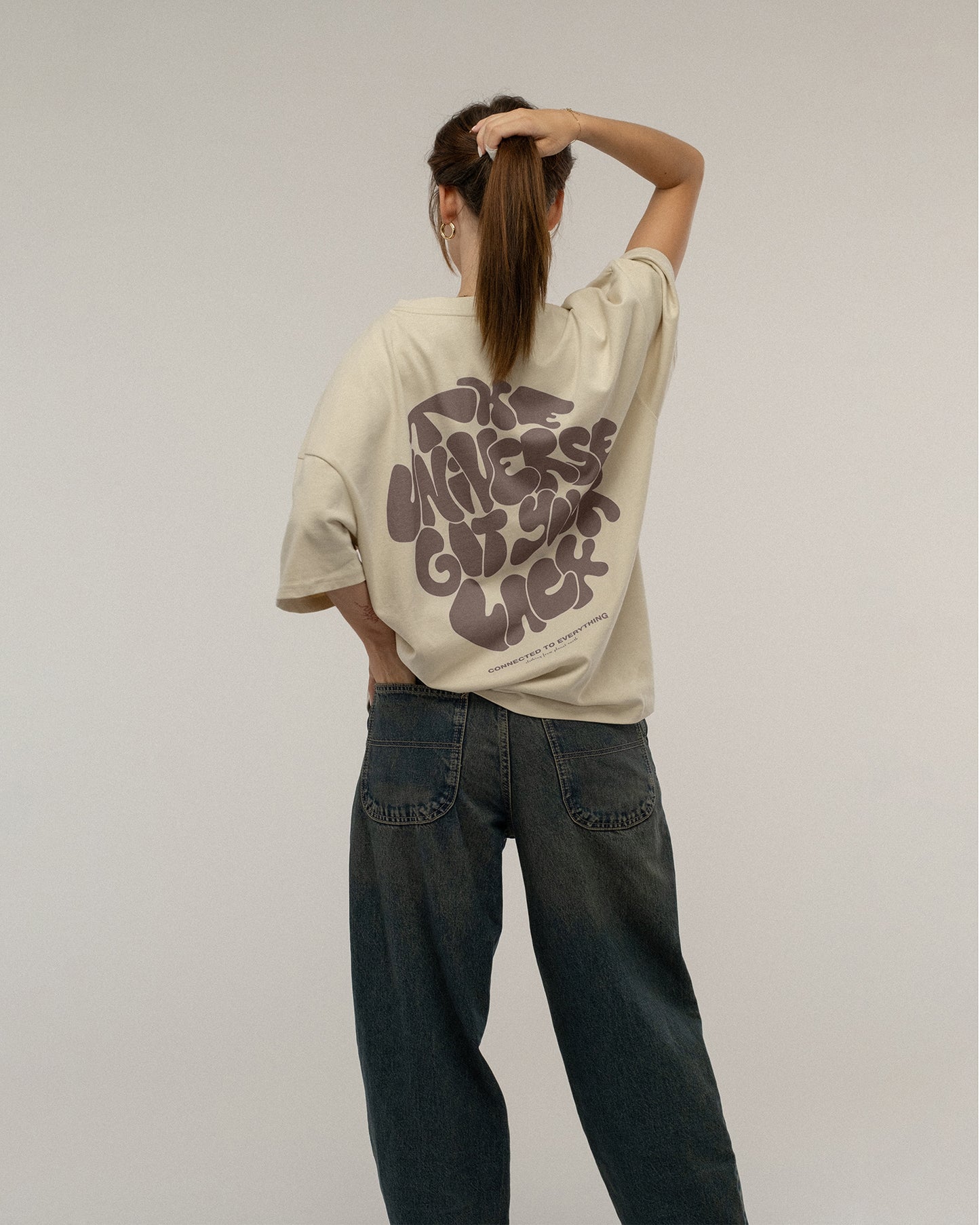 Beiges T-Shirt mit dunkelbraunem Rückendruck wird von hinten getragen. Die Trägerin greift sich in die Haare. Der gesamte Motivtext „THE UNIVERSE GOT YOUR BACK“ mit darunter stehendem Slogan ist sichtbar.