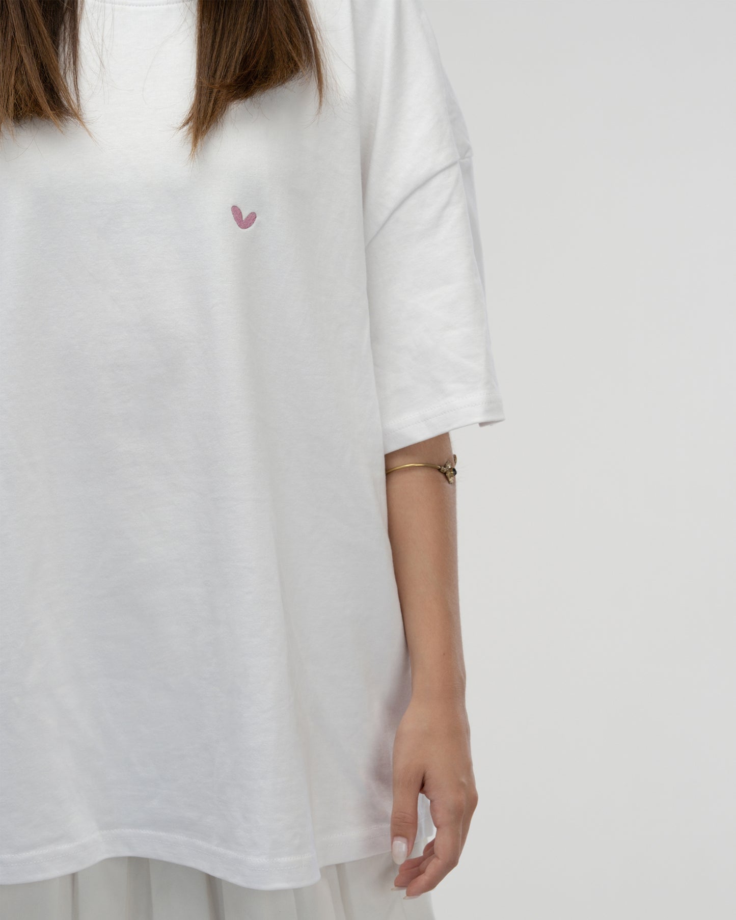 Nahaufnahme der Vorderseite eines weißen T-Shirts, das eine stehende Person trägt. Auf der linken Brust ist ein kleines, rosafarbenes, gesticktes Herzsymbol zu sehen.