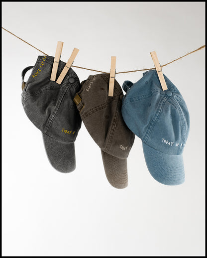 Drei Baseballcaps in Schwarz, Braun und Blau hängen mit Wäscheklammern an einer Leine; auf den Kappen sind Slogans wie „Today is a good day“ und „Made on planet Earth“ gestickt.