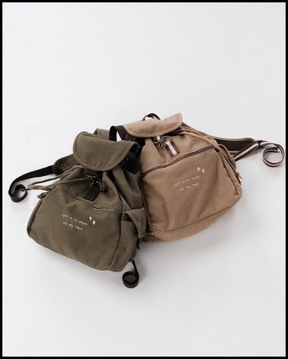 Zwei Canvas-Rucksäcke in gedeckten Farben liegen nebeneinander auf hellem Untergrund. Links befindet sich ein olivgrünes Modell, rechts ein beigefarbenes. Beide Rucksäcke besitzen eine Fronttasche mit Reißverschluss, eine Klappe mit Schnalle sowie seitliche Taschen. Auf der Front ist jeweils in heller Schrift der Spruch „LET’S GO TO WHERE WE ARE FREE“ mit kleinen Sternsymbolen aufgestickt.