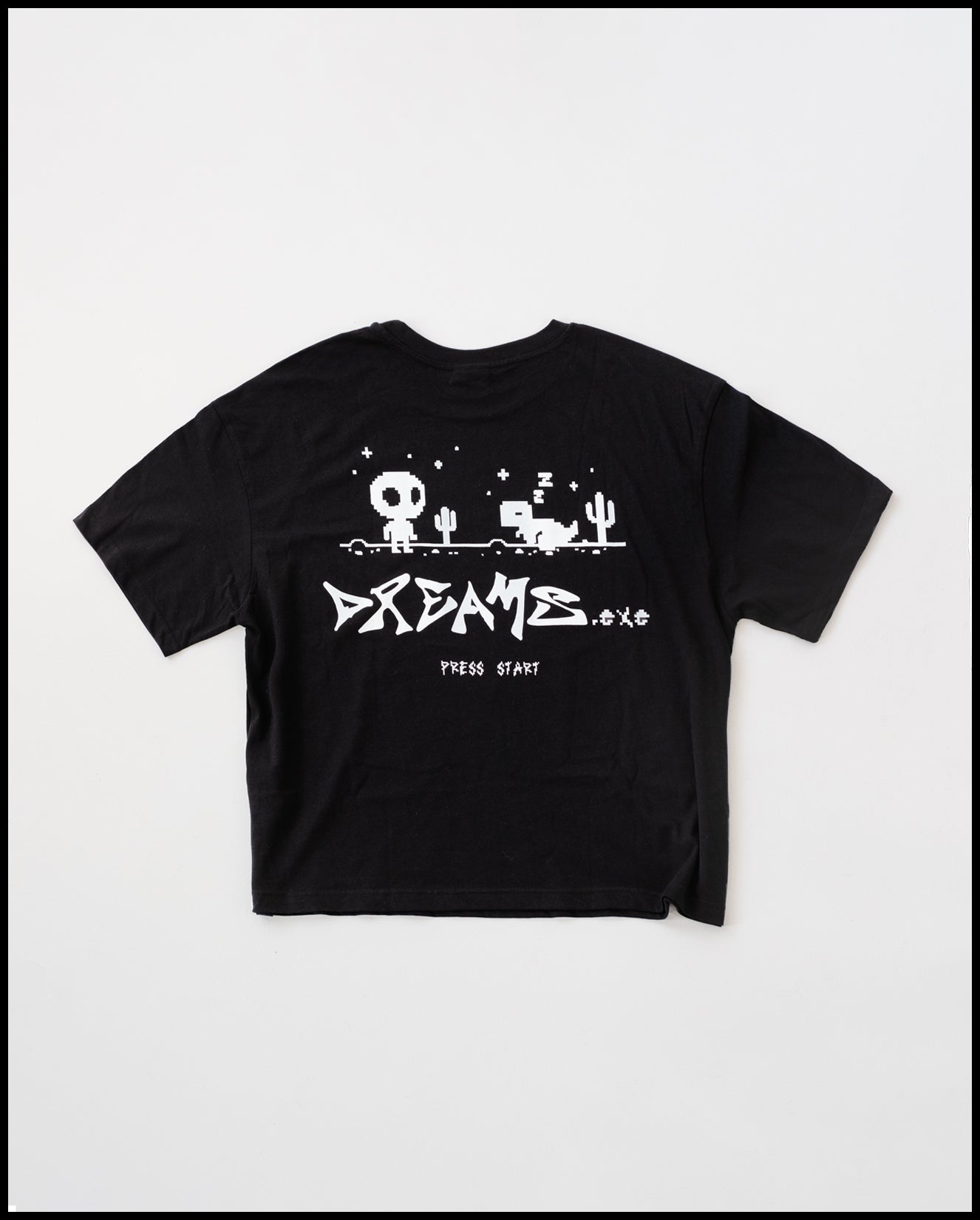 Schwarzes T-Shirt mit großem weißen Rückendruck im Pixel-Art-Stil. Das Motiv zeigt eine Spiellandschaft mit Kaktus, einem stehenden Figur-Charakter mit großem Kopf sowie einem schlafenden Pixel-Tier. Darunter steht in markanter Schrift „DREAMS.exe“ und klein „PRESS START“.