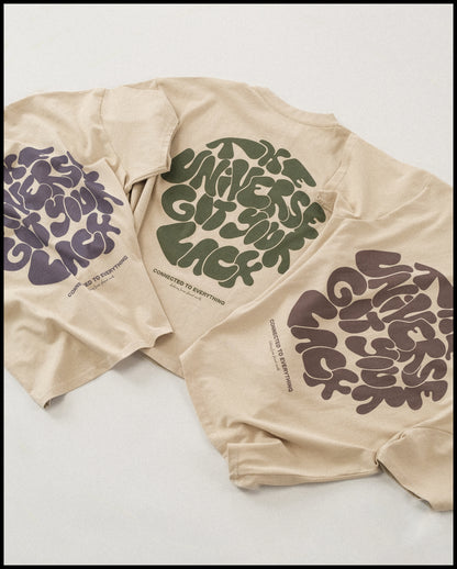 Drei beigefarbene T-Shirts liegen nebeneinander und zeigen jeweils einen großflächigen Rückendruck mit dem Schriftzug „THE UNIVERSE GOT YOUR BACK“ in organisch geschwungener Typografie. Die Druckfarben variieren in Dunkelviolett, Grün und Braun. Unterhalb des Motivs steht in kleiner Schrift „CONNECTED TO EVERYTHING – clothing from planet earth“.
