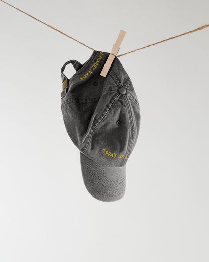 Schwarze Baseballcap hängt mit einer Wäscheklammer an einer Leine, Stickerei in Gelb auf Schirm und Seitenpanel mit dem Text „Today is a good day“ und „Planet Earth“.