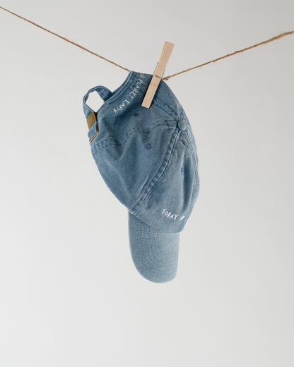 Blaue Baseballcap hängt an einer Leine, weiße Stickerei mit dem Text „Today is a good day“ und „Planet Earth“ sichtbar auf Schirm und Seite.