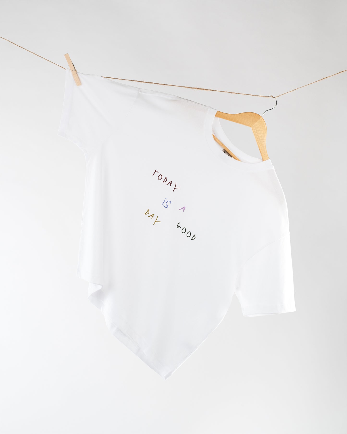  Weißes T-Shirt mit lockerem Schnitt hängt schräg auf einem Holzkleiderbügel an einer Wäscheleine; auf der Vorderseite befindet sich ein bunter, handschriftlicher Schriftzug „Today is a good day“.
