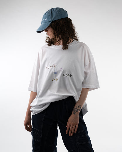 Person trägt das weiße T-Shirt mit bunter Stickerei „Today is a good day“, kombiniert mit einer blauen Cap und dunkler Cargohose, Blick ist zur Seite gerichtet.