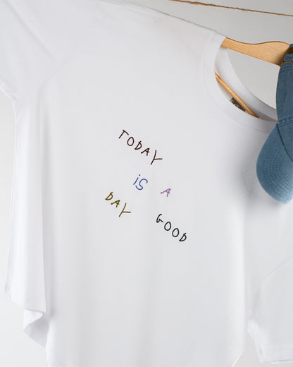 Detailaufnahme des weißen T-Shirts auf Kleiderbügel, Fokus auf den bunten Stick-Schriftzug „Today is a good day“, eine blaue Cap hängt ebenfalls am Bügel.
