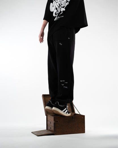 Person in schwarzer Jogginghose mit weißen Stickdetails auf der Rückseite: drei kleine Sterne und der Schriftzug ‚REACH FOR THE STARS..‘ auf der rechten Beinrückseite. Getragen mit weiß-schwarzen Sneakern und einem schwarzen Shirt mit großem weißen Print.