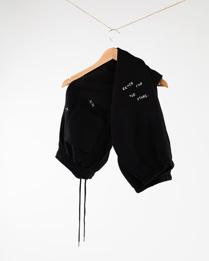Schwarze Jogginghose locker über einen Holzkleiderbügel gehängt, sichtbar ist der weiße Sticktext ‚REACH FOR THE STARS..‘ sowie zwei kleine gestickte Kreuze oberhalb der Tasche. Hosenbeine mit Gummizug und Tunnelzug
