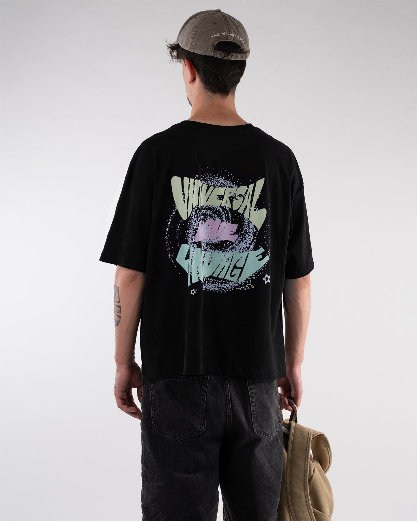 Person mit schwarzem T-Shirt in Rückansicht. Der große Rückendruck zeigt pastellfarbeneSchrift umgeben von einem gesprenkelten Kreis mit dem Schriftzug ‚UNIVERSAL LOVE LANGUAGE‘ in verspielter Typografie. Die Person trägt eine graue Cap und hält einen beigefarbenen Rucksack.