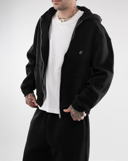 Person mit geöffnetem, schwarzem Zip-Hoodie über einem weißen T-Shirt. Auf der linken Brust des Hoodies ist ein kleines, weißes Peace-Handzeichen aufgestickt. Die Person trägt eine schwarze Jogginghose und hat sichtbare Tattoos an Hals und Händen.