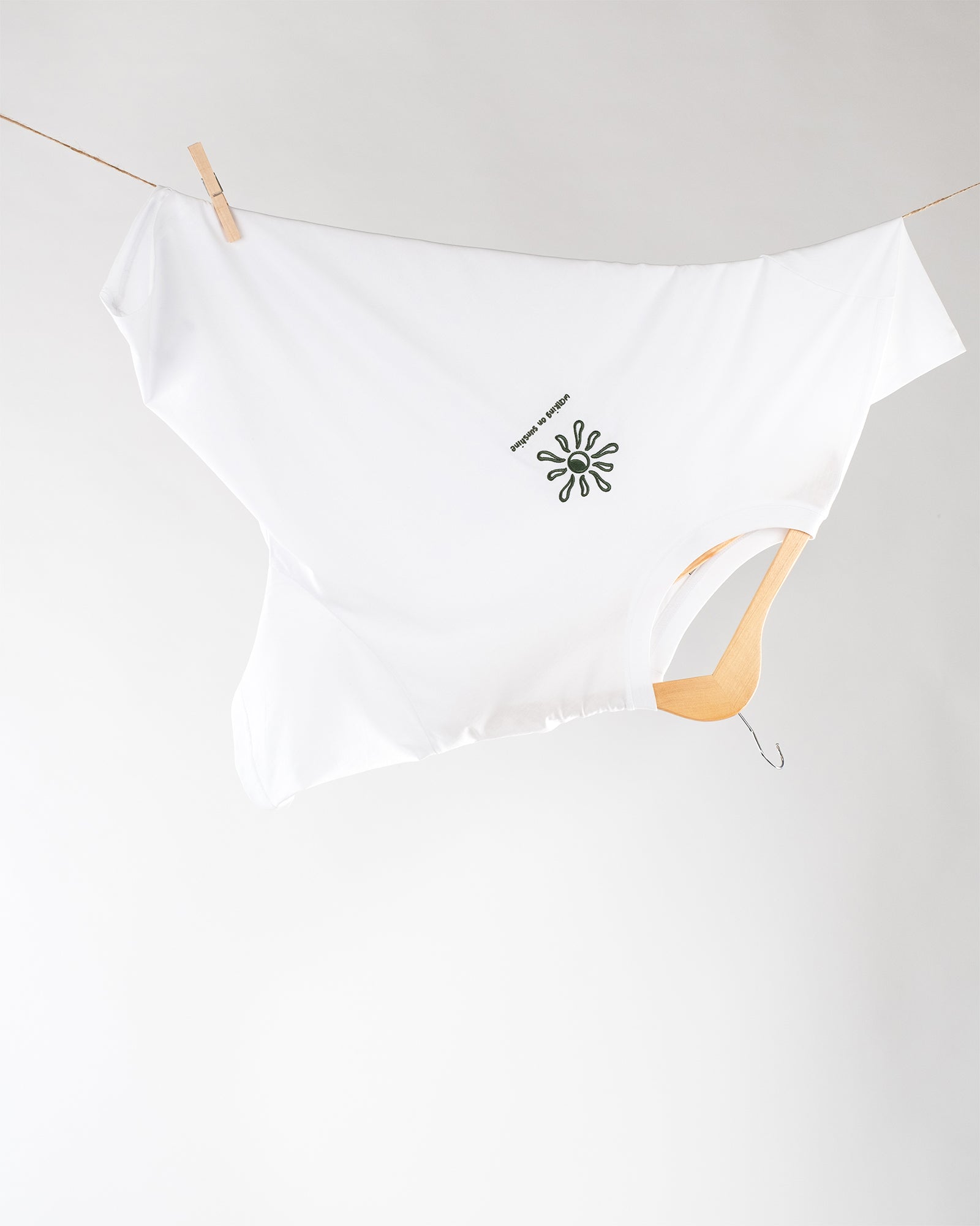 Weißes T-Shirt, aufgehängt auf einem Holzkleiderbügel und einer Wäscheleine, seitlich fixiert mit einer Holzklammer. In der Mitte des T-Shirts befinmdet sich eine Sonnenform mit dem Text ‚WALKING ON SUNSHINE‘.