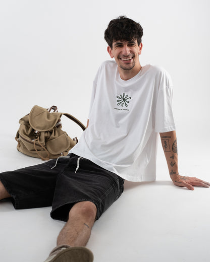 Lächelnde Person sitzt entspannt auf dem Boden und trägt ein weißes T-Shirt mit grünem Sonnensymbol und dem Schriftzug ‚walking on sunshine‘. Dazu schwarze Shorts mit Kordelzug. Im Hintergrund liegt ein beiger Rucksack. Sichtbare Tattoos an Armen und Beinen
