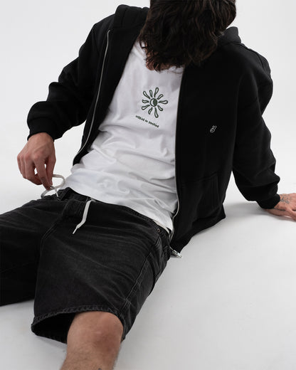 Sitzende Person in schwarzem Zip-Hoodie, weißem T-Shirt mit grün gestickter Sonne und dem Text ‚walking on sunshine‘, sowie schwarzer Shorts mit Kordelzug. Der Hoodie ist offen getragen und zeigt ein kleines weißes Peace-Zeichen auf der linken Brust.