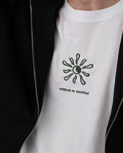 Nahaufnahme eines weißen T-Shirts mit grüner Stickerei auf der Brust: grafische Sonnendarstellung und der Text ‚walking on sunshine‘ in Kleinbuchstaben. Darüber wird ein schwarzer Zip-Hoodie offen getragen.