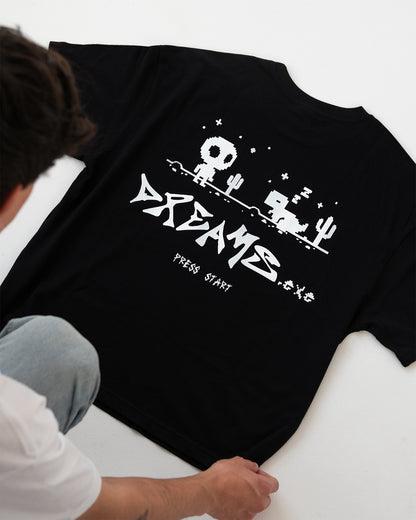 T-Shirt im Flatlay, Rückseite oben: kompletter weißer Pixel-Rückenprint sichtbar – Totenkopf-Figur, Kaktus, schlafendes Tier, Sterne – mit großem „DREAMS“-Schriftzug und „PRESS START“ darunter.