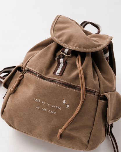 Detailaufnahme sandfarbener Canvas-Rucksack; Überschlag mit Metallhaken und braun-weißem Band, Front-Reißverschluss, Kordelenden; weiße Stickerei „LET’S GO TO WHERE WE ARE FREE“.