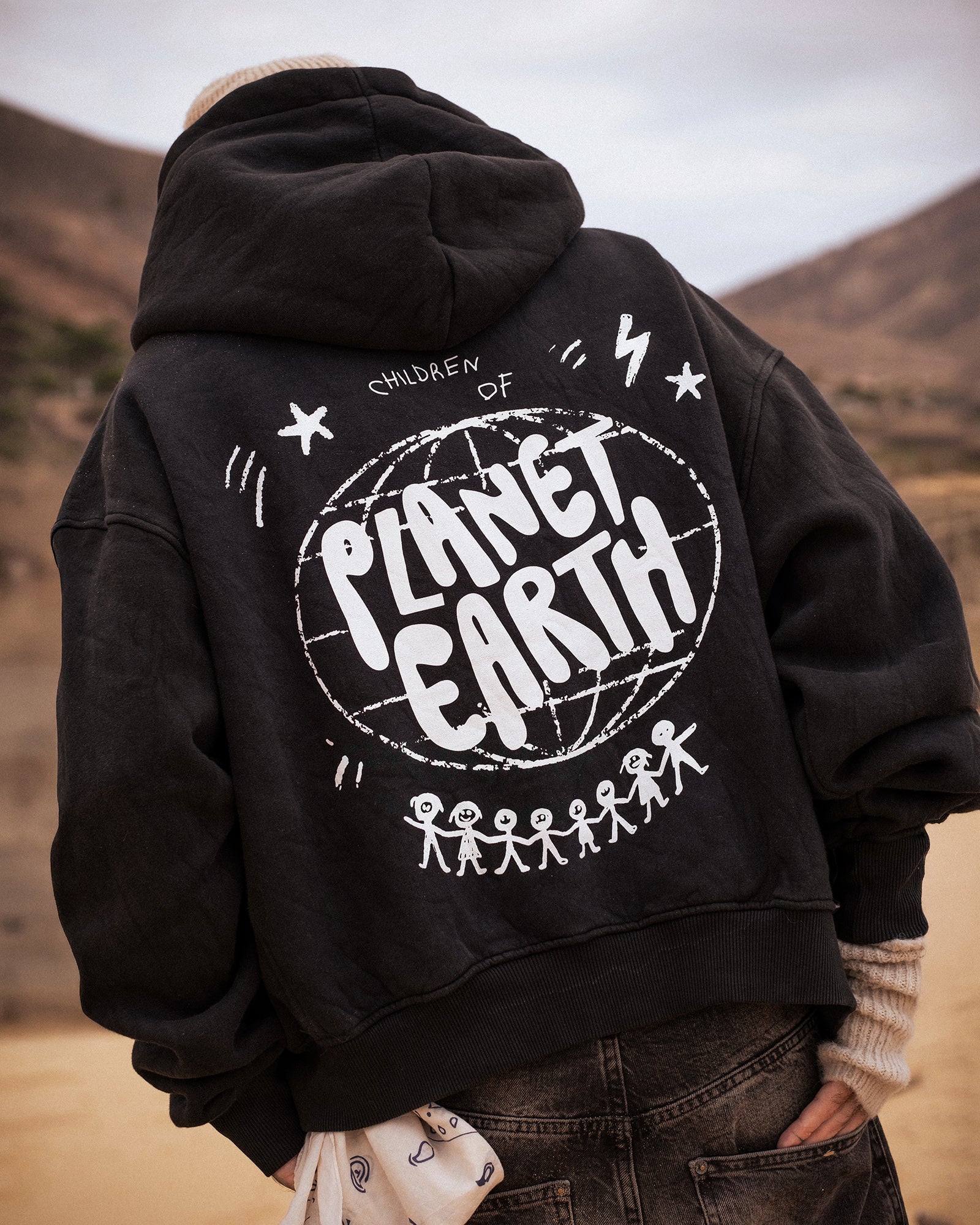 Person trägt eine schwarze Oversized-Hoodie-Jacke mit großer weißer Rückengrafik. Das Design zeigt den Schriftzug „Children of Planet Earth“ mit einer Weltkugel und stilisierten Strichmännchen, die sich an den Händen halten. Der Stil ist lässig und urban, ideal für Streetwear-Looks