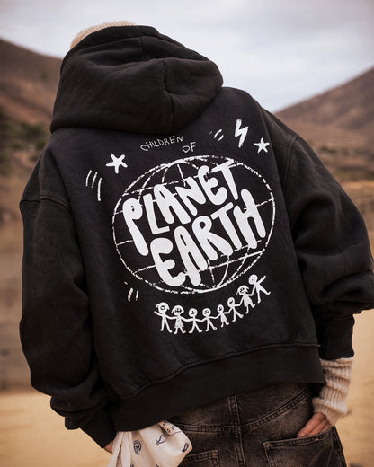 Person trägt eine schwarze Oversized-Hoodie-Jacke mit großer weißer Rückengrafik. Das Design zeigt den Schriftzug „Children of Planet Earth“ mit einer Weltkugel und stilisierten Strichmännchen, die sich an den Händen halten. Der Stil ist lässig und urban, ideal für Streetwear-Looks