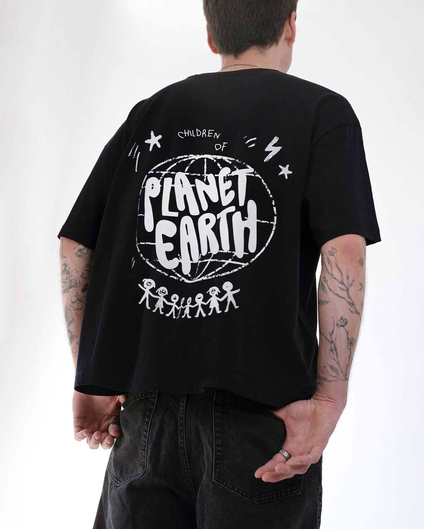 Person trägt ein schwarzes Oversized-T-Shirt mit großem weißen Rückendruck „Children of Planet Earth“. Das Design zeigt eine Weltkugel, Sterne und Strichmännchen, die sich an den Händen halten. Lässiger Streetwear-Stil mit nachhaltiger Botschaft, kombiniert mit schwarzen Jeans.
