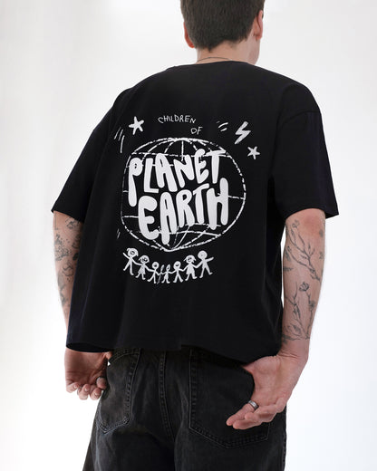 Person trägt ein schwarzes Oversized-T-Shirt mit großem weißen Rückendruck „Children of Planet Earth“. Das Design zeigt eine Weltkugel, Sterne und Strichmännchen, die sich an den Händen halten. Lässiger Streetwear-Stil mit nachhaltiger Botschaft, kombiniert mit schwarzen Jeans.