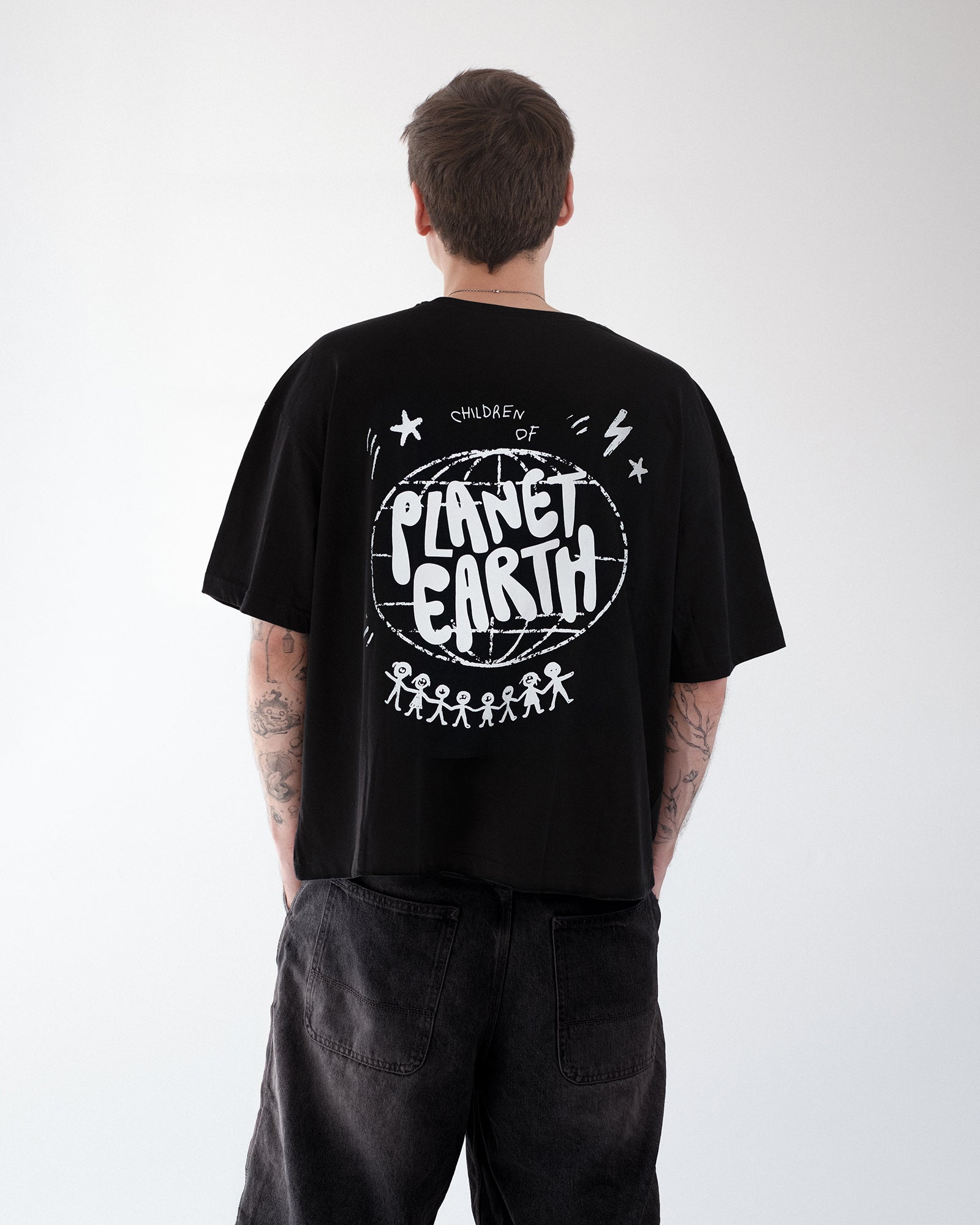 Rückansicht einer Person mit schwarzem Oversized-T-Shirt, bedruckt mit dem weißen Motiv „Children of Planet Earth“. Das Design zeigt eine Weltkugel, Sterne und eine Reihe von Strichmännchen, die sich an den Händen halten. Moderner Streetwear-Look, kombiniert mit locker sitzender schwarzer Jeans.