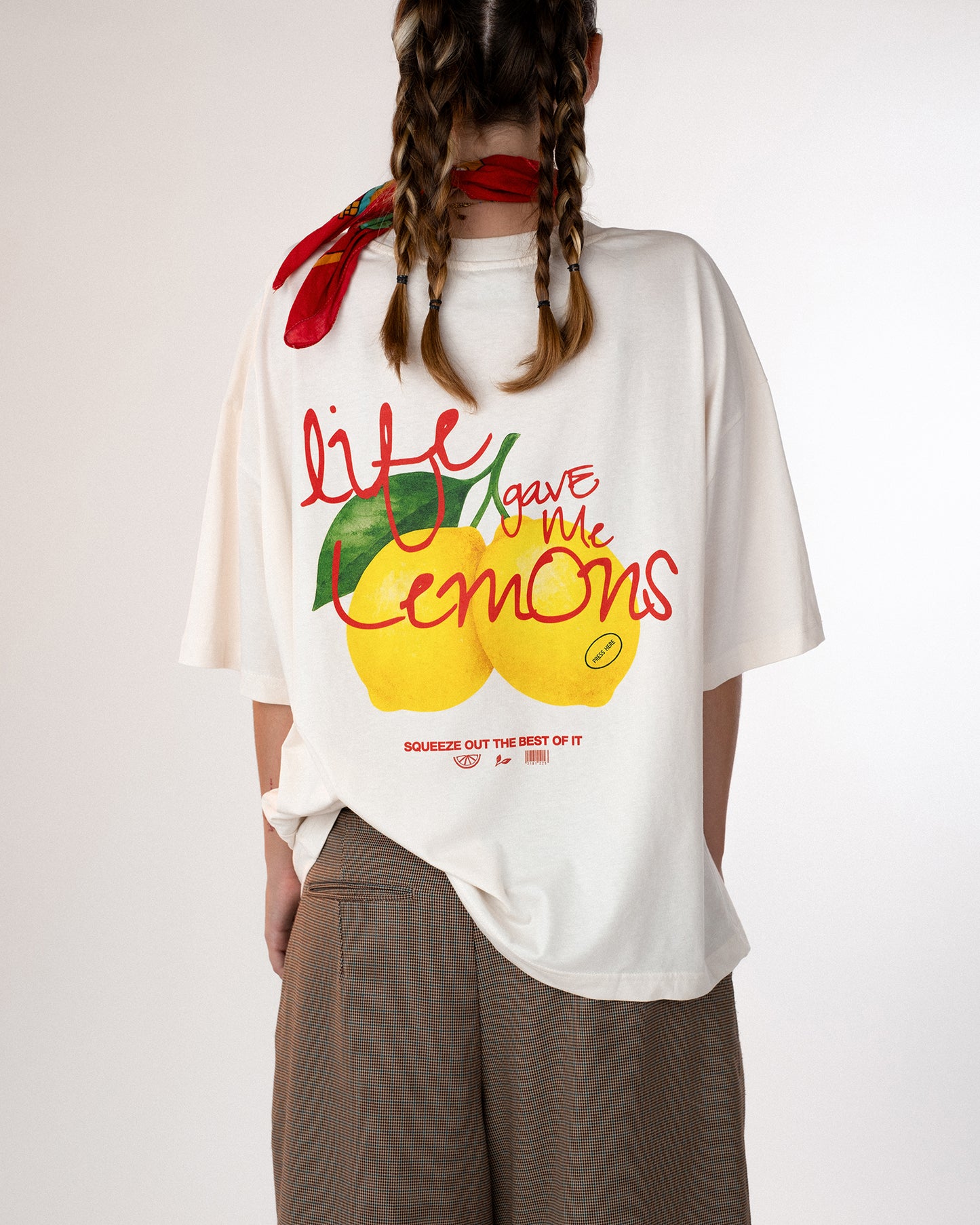 Rückansicht einer Person mit weißem Oversized-T-Shirt, bedruckt mit buntem Zitronenmotiv und dem Schriftzug „Life gave me lemons – squeeze out the best of it“. Kombiniert mit braunen Stoffshorts und rotem Halstuch – frischer, sommerlicher Streetwear-Look mit positiver Botschaft.Botschaft.