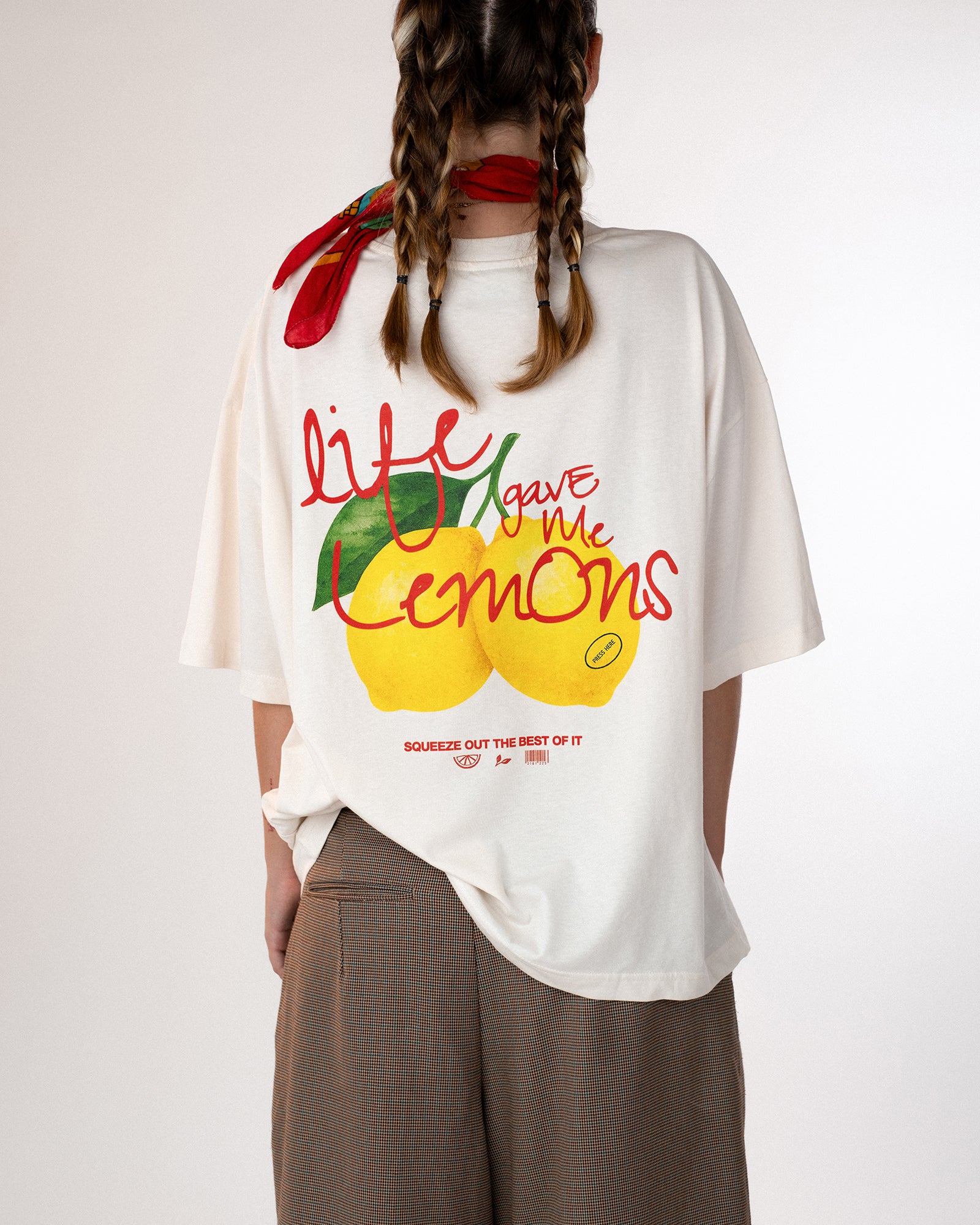 Rückansicht einer Person mit weißem Oversized-T-Shirt, bedruckt mit buntem Zitronenmotiv und dem Schriftzug „Life gave me lemons – squeeze out the best of it“. Kombiniert mit braunen Stoffshorts und rotem Halstuch – frischer, sommerlicher Streetwear-Look mit positiver Botschaft.Botschaft.