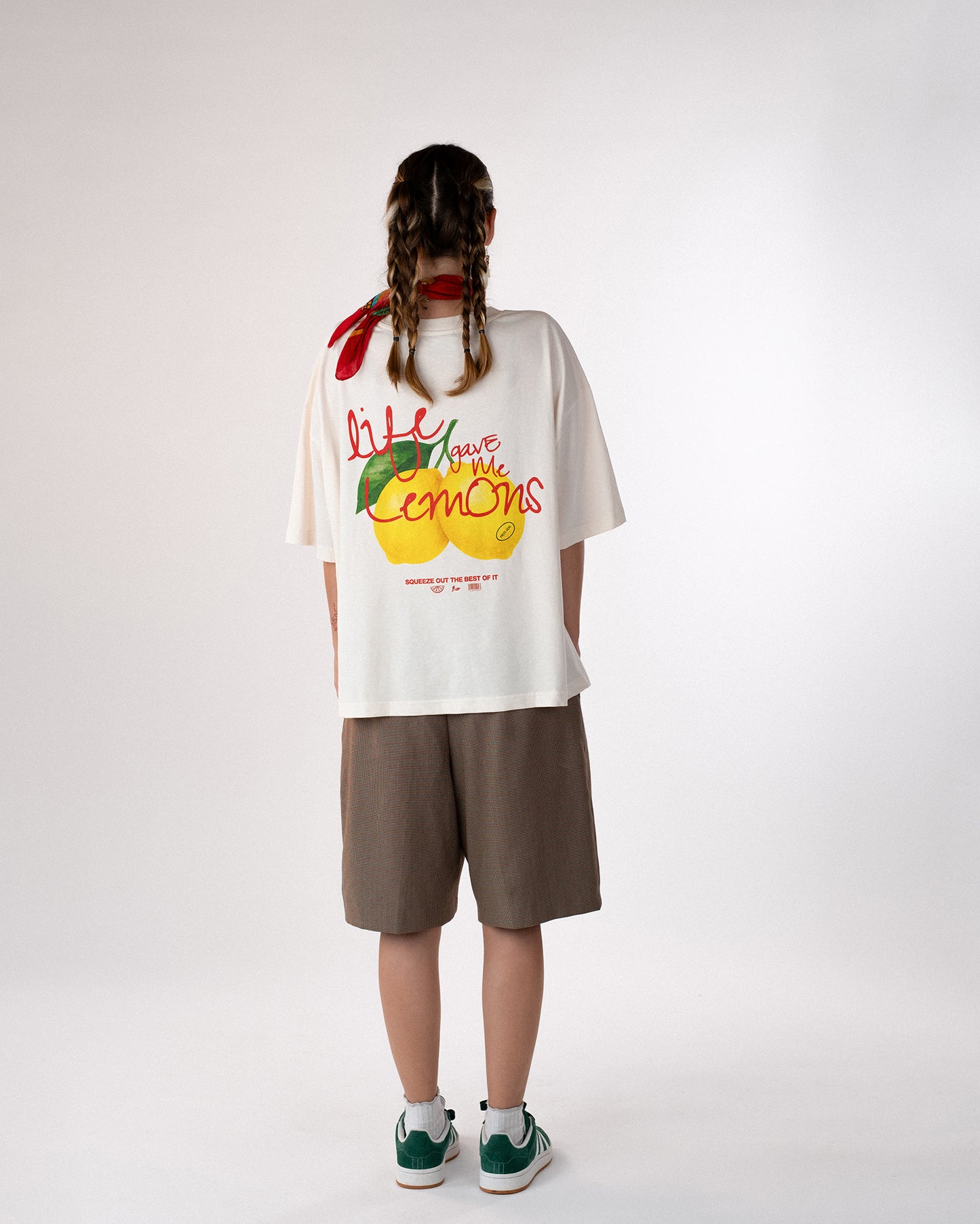Person steht mit dem Rücken zur Kamera und trägt ein weißes Oversized-T-Shirt mit buntem Rückendruck „Life gave me lemons – squeeze out the best of it“. Kombiniert mit braunen Shorts, grünen Sneakern und rotem Halstuch – lässiger, sommerlicher Streetwear-Look mit positiver Botschaft.