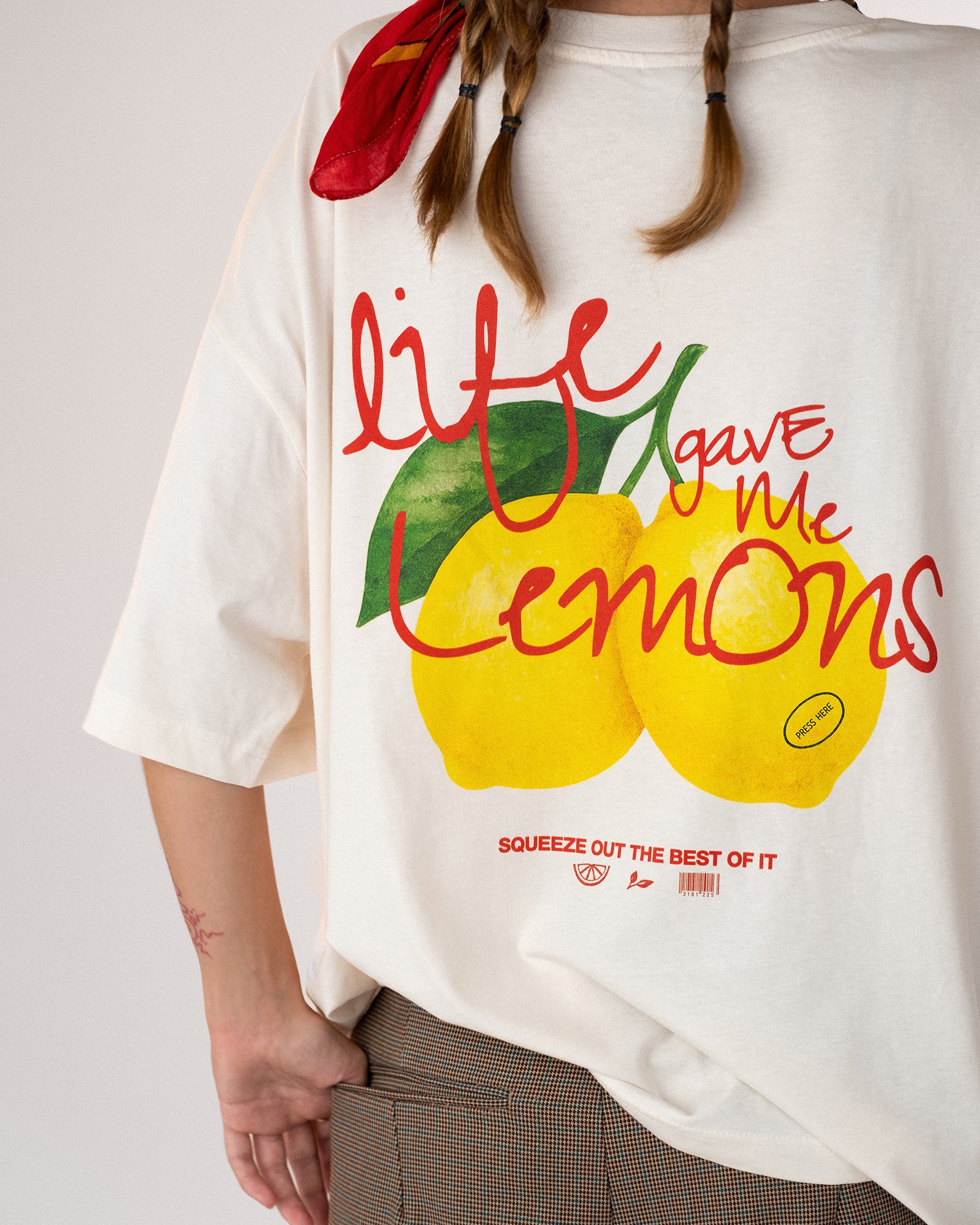 Nahaufnahme eines weißen Oversized-T-Shirts mit buntem Zitronen-Print und dem Schriftzug „Life gave me lemons – squeeze out the best of it“. Frisches, sommerliches Design mit handgeschriebener Typografie in Rot und Grün, getragen zu braunen Shorts – moderner Streetwear-Look mit positiver Message.
