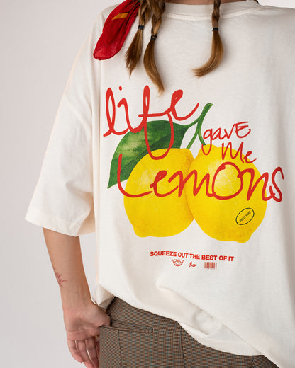 Nahaufnahme eines weißen Oversized-T-Shirts mit buntem Zitronen-Print und dem Schriftzug „Life gave me lemons – squeeze out the best of it“. Frisches, sommerliches Design mit handgeschriebener Typografie in Rot und Grün, getragen zu braunen Shorts – moderner Streetwear-Look mit positiver Message.