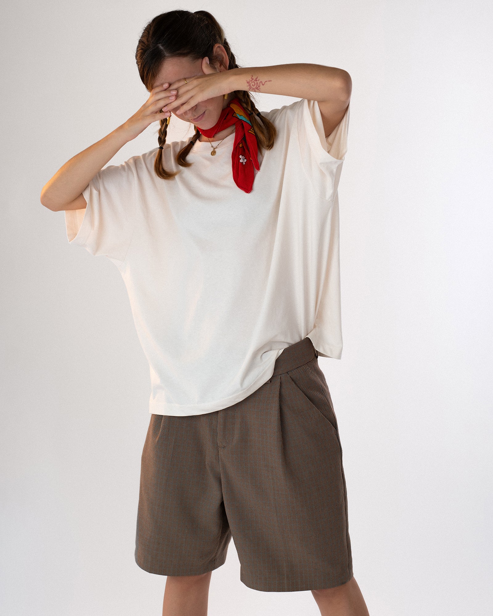 Person trägt ein weißes Oversized-T-Shirt ohne sichtbaren Aufdruck auf der Vorderseite, kombiniert mit braunen Shorts und einem roten Halstuch. Lässiger, sommerlicher Look mit verspielter Pose – moderne Streetwear mit minimalistischer Ästhetik.