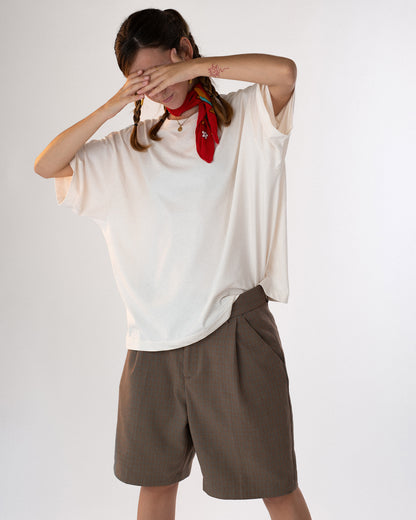 Person trägt ein weißes Oversized-T-Shirt ohne sichtbaren Aufdruck auf der Vorderseite, kombiniert mit braunen Shorts und einem roten Halstuch. Lässiger, sommerlicher Look mit verspielter Pose – moderne Streetwear mit minimalistischer Ästhetik.