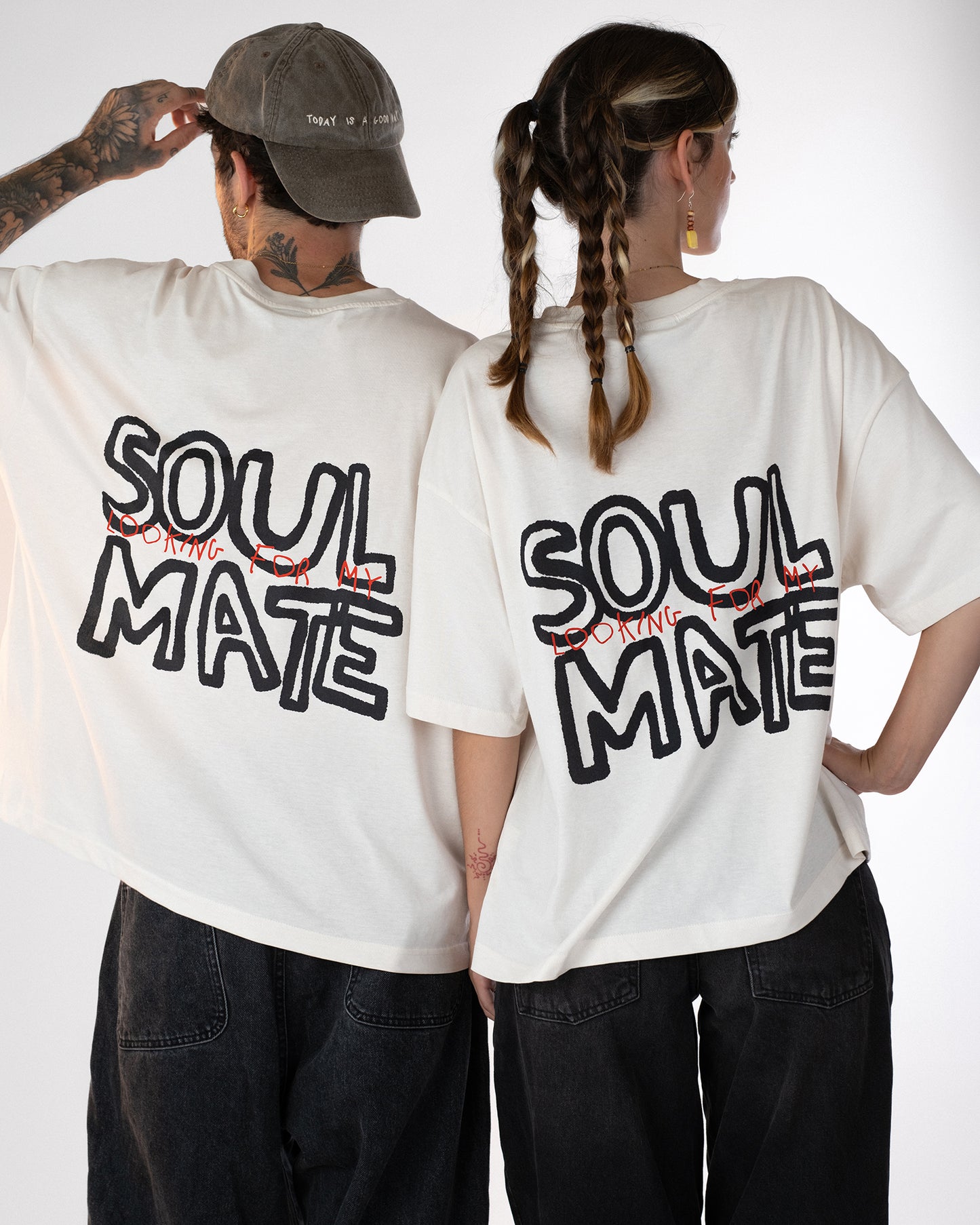 Zwei Personen stehen Rücken an Rücken und tragen weiße Oversized-T-Shirts mit schwarzem und rotem Rückendruck „Looking for my Soul Mate“. Beide Outfits sind im lässigen Unisex-Streetwear-Stil mit dunklen Jeans gestaltet – modernes Partner-Look-Shirt mit verspielter Botschaft.
