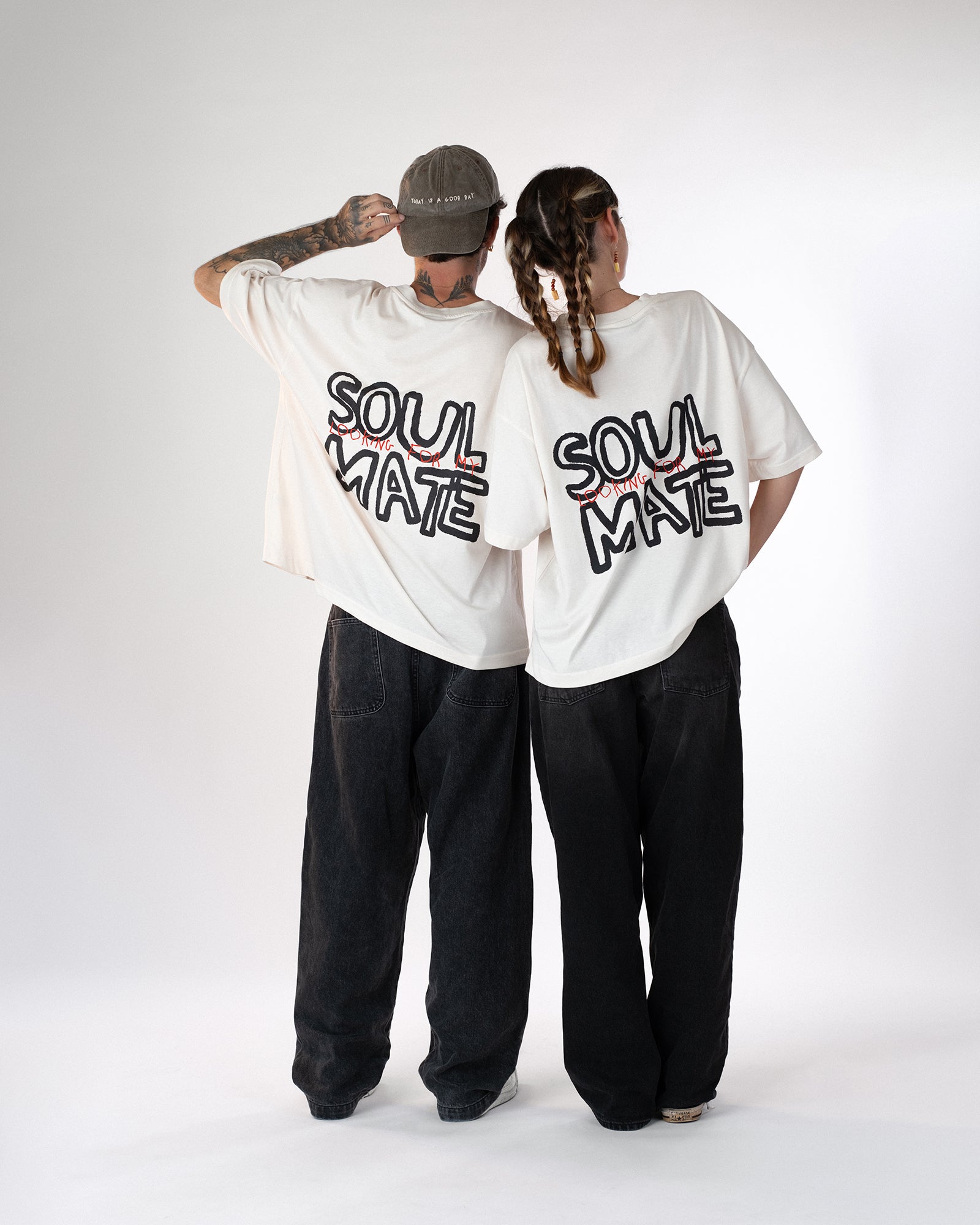 Zwei Personen stehen nebeneinander und tragen weiße Oversized-T-Shirts mit schwarzem und rotem Rückendruck „Looking for my Soul Mate“. Beide kombinieren die Shirts mit weiten schwarzen Jeans – moderner Unisex-Partnerlook im entspannten Streetwear-Stil.