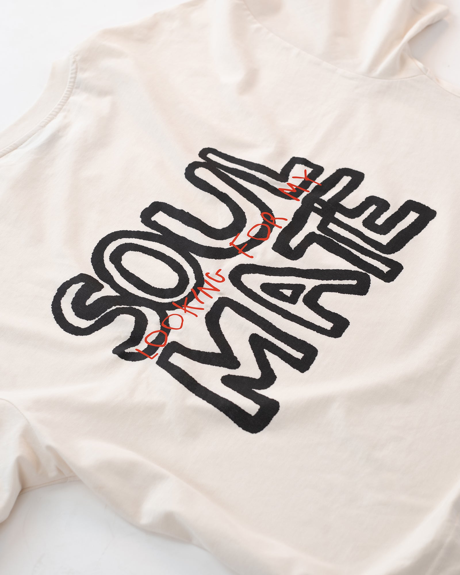 Detailaufnahme eines weißen T-Shirts mit großem schwarzen und roten Rückendruck „Looking for my Soul Mate“. Das Shirt liegt flach auf hellem Hintergrund – modernes Unisex-Design mit auffälliger Typografie im Streetwear-Stil.