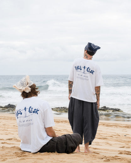 Zwei Personen am Strand tragen weiße Oversized-T-Shirts mit blauem Rückendruck „Take a Risk – To Be Brave“. Eine Person sitzt im Sand, die andere steht, beide mit Bandanas und weiten Hosen. Entspannter, inspirierender Beachwear-Look im modernen Streetwear-Stil.