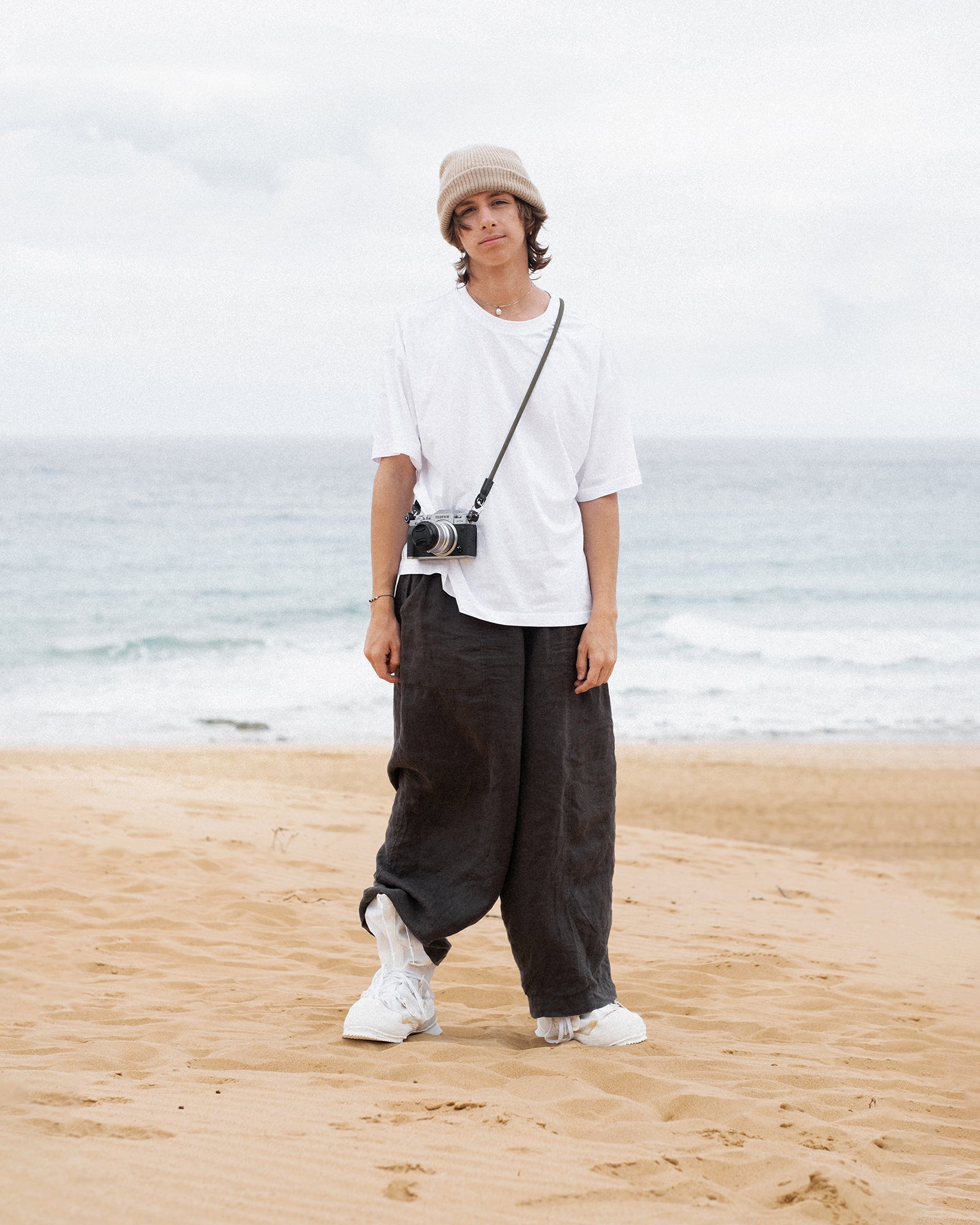 Person steht am Strand und trägt ein schlichtes weißes Oversized-T-Shirt, weite dunkle Hosen und weiße Sneaker. Ergänzt wird der Look durch eine beige Strickmütze und eine umgehängte Kamera – entspannter, minimalistischer Streetwear-Stil mit künstlerischem Touch.