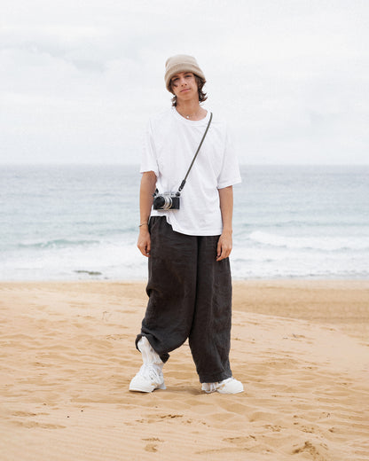 Person steht am Strand und trägt ein schlichtes weißes Oversized-T-Shirt, weite dunkle Hosen und weiße Sneaker. Ergänzt wird der Look durch eine beige Strickmütze und eine umgehängte Kamera – entspannter, minimalistischer Streetwear-Stil mit künstlerischem Touch.