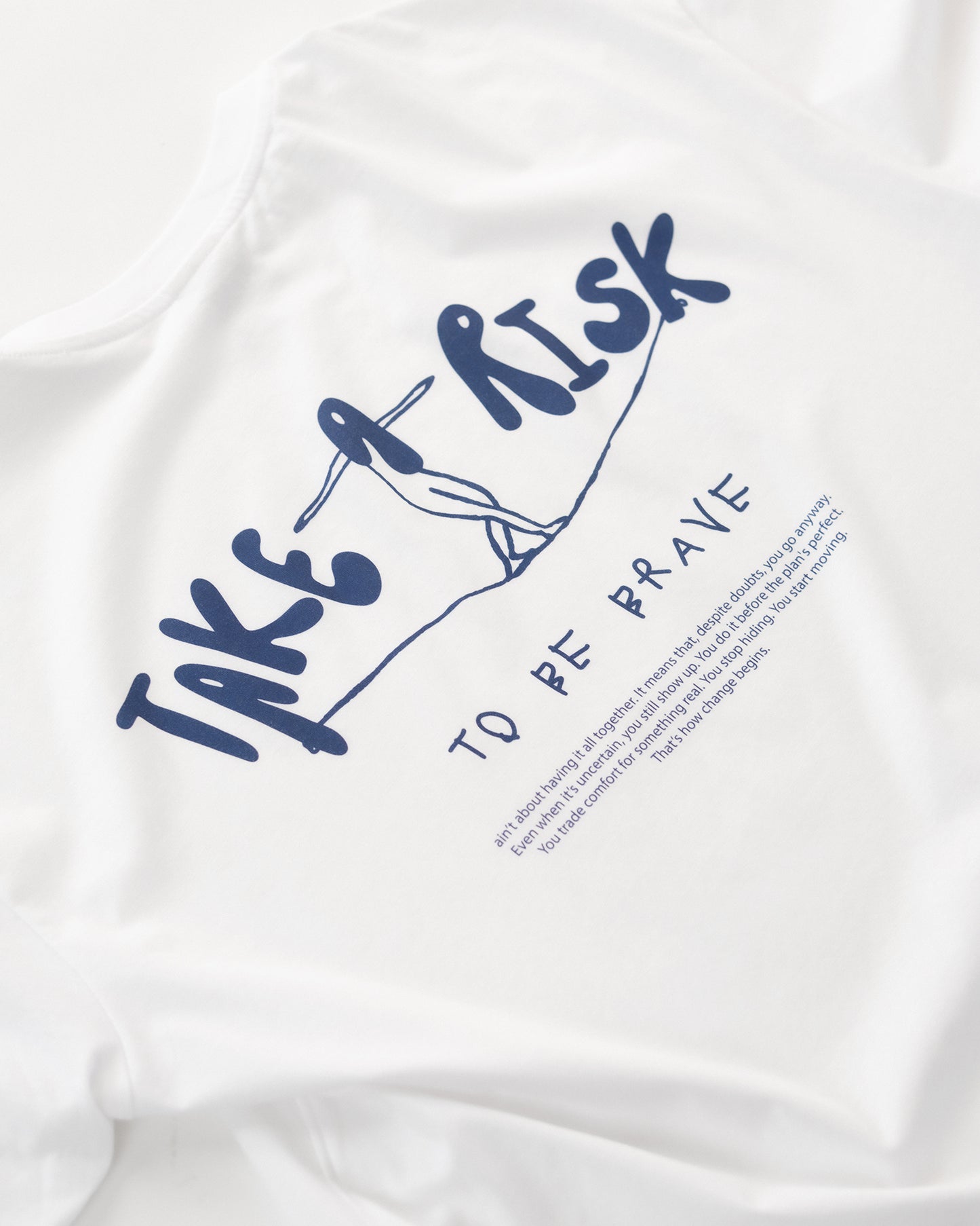 Weißes T-Shirt mit blauem Rückendruck „Take a Risk – To Be Brave“. Das Design zeigt eine stilisierte Figur auf einem Seil und darunter einen motivierenden Text – minimalistisches, inspirierendes Streetwear-Design, flach auf hellem Hintergrund präsentiert.