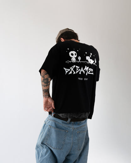 Schwarzes Oversize-T-Shirt mit großem Rückenprint in Weiß: Pixel-Wüstenmotiv mit Figur im Totenkopf-Stil, Kaktus und schlafendem Tier („Z“), darunter der Schriftzug „DREAMS“ und klein „PRESS START“; lockerer, weiter Schnitt.