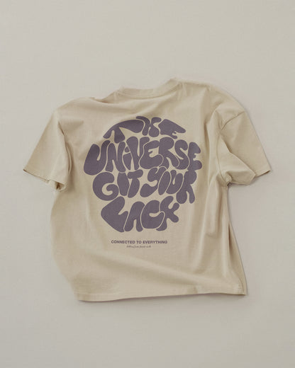 Beigefarbenes T-Shirt liegt flach auf hellem Untergrund. Auf dem Rücken ist in großer, lila, verspielter Typografie der Text „THE UNIVERSE GOT YOUR BACK“ zu sehen. Darunter steht klein „CONNECTED TO EVERYTHING – clothing from planet earth“.