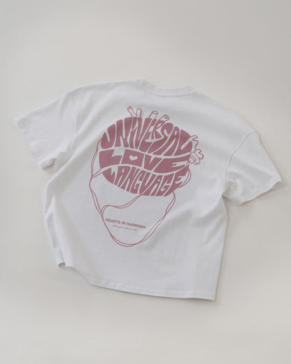 Weißes T-Shirt liegt flach mit dem Rücken nach oben. Auf dem Rücken ist ein großes, herzförmiges Motiv in Rosa zu sehen, das aus den Worten „UNIVERSAL LOVE LANGUAGE“ besteht. Darunter steht in kleiner Schrift: „HEARTS IN HARMONY – clothing from planet earth“.