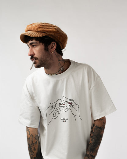 Weißes T-Shirt mit schwarzer Liniengrafik von zwei Händen, die gemeinsam einen mit Herzen verzierten Joint drehen. Darunter steht in kleiner Schrift: „inhale my love“.