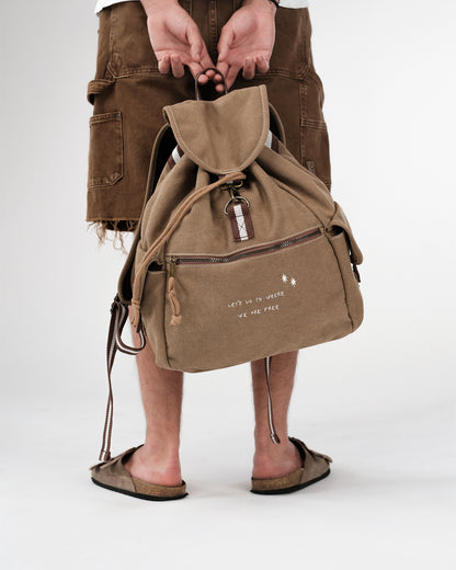 Sandfarbener Canvas-Rucksack, Kordelzug unter Überschlag, mittiger Metallhaken, Front-Reißverschlusstasche, seitliche Pattentaschen; weiße Stickerei „LET’S GO TO WHERE WE ARE FREE“.
